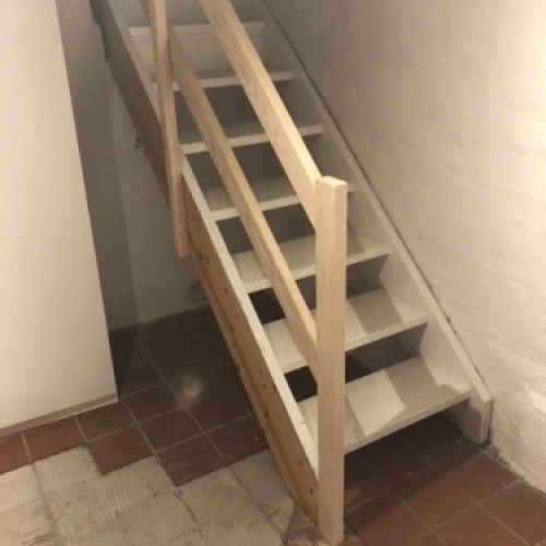 Et moderne gelænder i lyst træ og hvide balustre, der løber langs en trappe i et lyst og åbent hjem.