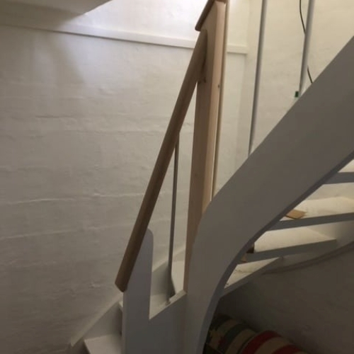 Et lyst gelænder monteret på en hvidmalet trappe med kurvet form, hvor træet står i kontrast til de hvide vægge.