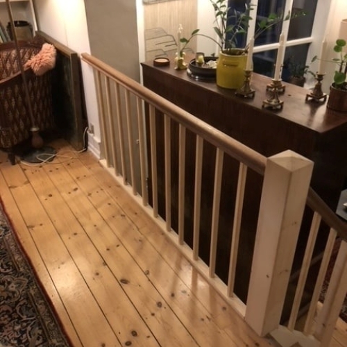 Et elegant gelænder i lyst træ og mørk håndliste, placeret i en stue med grøn sofa og store vinduer.