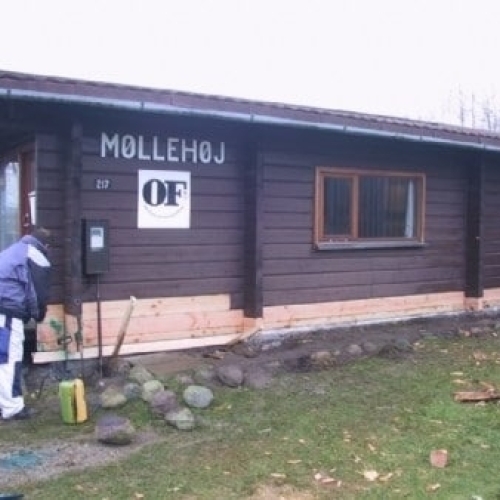 En person arbejder på at male eller renovere et mørkebrunt træhus med skiltet 'MØLLEHØJ' på facaden. Der er brædder udskiftet langs husets sokkel og byggematerialer ligger spredt på jorden.