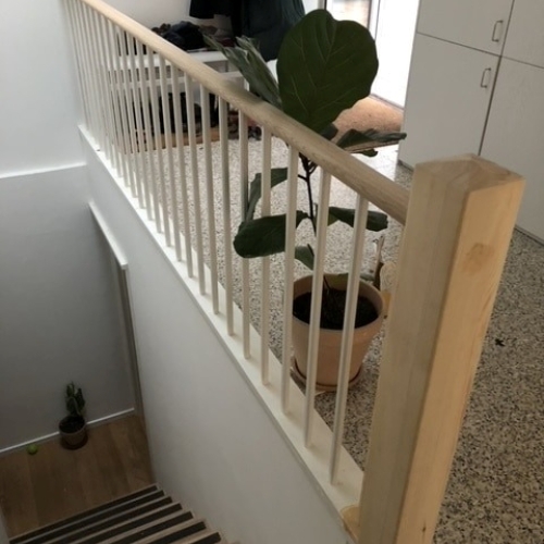 Et langt gelænder i lyst træ med lodrette spiler, placeret langs repos i en hyggelig stue med planter og lamper.