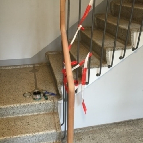 Et gelænder i træ og stål i en opgang, under reparation, med rødt og hvidt tape omkring håndlisten.