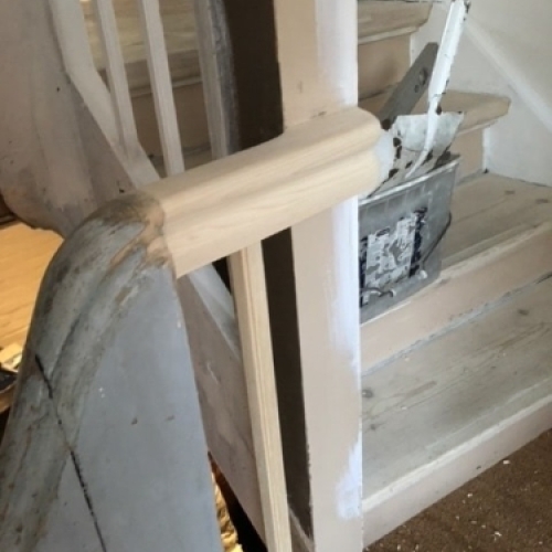 Et gelænder i lyst træ med lodrette spiler, placeret på første sal ved en lys trappe ned mod kælderen.