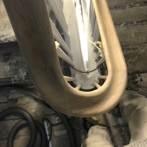 Et buet gelænder i lyst træ, fotograferet ovenfra under montering på en trappe med hvide balustre.