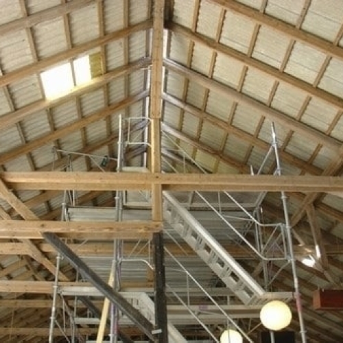 Indvendig udsigt af en lade med synlige træbjælker og stillads under taget, hvor et ovenlysvindue i tagfladen lukker naturligt lys ind.