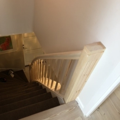 En tæppebelagt trappe med et robust gelænder i lyst træ og en solid firkantet stolpe i bunden.