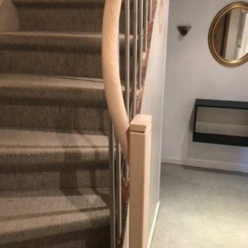 En stilren trappe med naturtræstrin, lodrette stålstænger og et elegant, mørkt gelænder langs væggen.