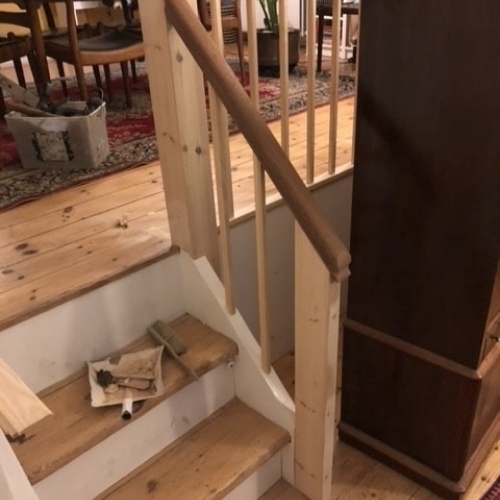 Et nybygget gelænder i lyst træ med mørk håndliste ved en trappe med fyrretræstrin, placeret i en stue med trægulv og tæppe.