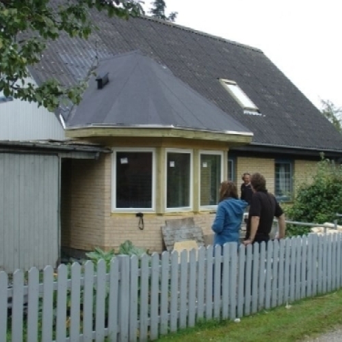 Tre personer står foran et hus med nyinstallerede vinduer og en grå træhegn, hvor der er i gang med udskiftning af døre og vinduer.